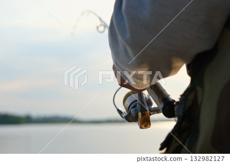 Old man fishing. Senior gray haired fisherman throws a spinning from shoreside at sunset, twisting a coil. Positive elderly male angling at lake, rotating reel rod. Retired fisher enjoying his weekend 132982127