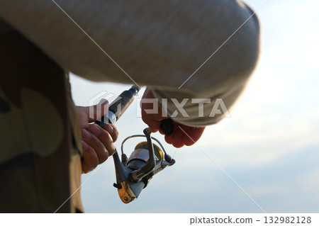 Old man fishing. Senior gray haired fisherman throws a spinning from shoreside at sunset, twisting a coil. Positive elderly male angling at lake, rotating reel rod. Retired fisher enjoying his weekend 132982128