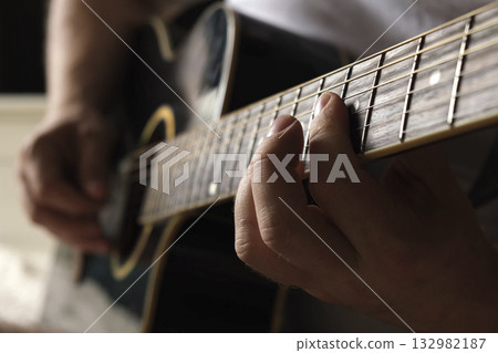 Playing the guitar. Strumming acoustic guitar. Musician plays music. Man fingers holding mediator. Man hand playing guitar neck in dark room. Unrecognizable person rehearsing, fretboard close-up Playing the guitar. Strumming acoustic guitar. Musician plays music. Man fingers holding mediator. Man hand playing guitar neck in dark room. Unrecognizable person rehearsing, fretboard close-up 132982187
