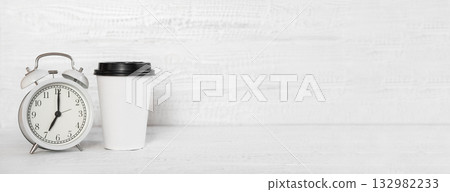 Front view white desk with alarm clock, coffee paper cup, copy space. Morning background banner with free space for text. Minimalistic concept workspace, time to work. Blank workplace. Office mock-up 132982233