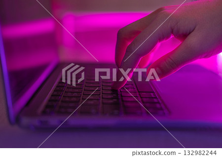 Person typing at laptop computer at night. Female uses keyboard, shakes buttons with fingers. Internet security, cyber attack. Trendy neon light. Creative vivid color of ultraviolet red and blue 132982244
