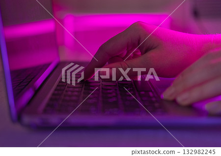 Person typing at laptop computer at night. Female uses keyboard, shakes buttons with fingers. Internet security, cyber attack. Trendy neon light. Creative vivid color of ultraviolet red and blue 132982245