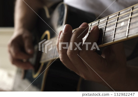 Playing the guitar. Strumming acoustic guitar. Musician plays music. Man fingers holding mediator. Man hand playing guitar neck in dark room. Unrecognizable person rehearsing, fretboard close-up Playing the guitar. Strumming acoustic guitar. Musician plays music. Man fingers holding mediator. Man hand playing guitar neck in dark room. Unrecognizable person rehearsing, fretboard close-up 132982247