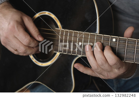 Playing the guitar. Strumming acoustic guitar. Musician plays music. Man fingers holding mediator. Man hand playing guitar neck in dark room. Unrecognizable person rehearsing, fretboard close-up 132982248