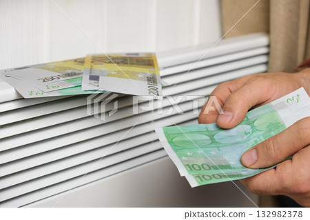 Human Hand Holding Euro Banknotes near Electric Heater Radiator at Home. Man Pay Money for Heating. Expensive to Heat. Energy Saving. Rising Cost of Living. Global Gas Crisis. Paying Cold Season Bills 132982378
