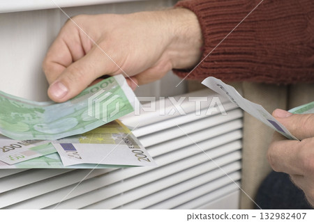 Human Hand Holding Euro Banknotes near Electric Heater Radiator at Home. Man Pay Money for Heating. Expensive to Heat. Energy Saving. Rising Cost of Living. Global Gas Crisis. Paying Cold Season Bills Human Hand Holding Euro Banknotes near Electric Heater Radiator at Home. Man Pay Money for Heating. Expensive to Heat. Energy Saving. Rising Cost of Living. Global Gas Crisis. Paying Cold Season Bills 132982407