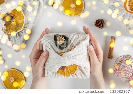 Gift Box in White Fabric. Female Hands wrapping Sustainable Gift on white Table with Decoration Elements. Dried Orange, Cinnamon, Cones. Zero waste Christmas Packaging. Hands decorating Furoshiki Gift 132982567
