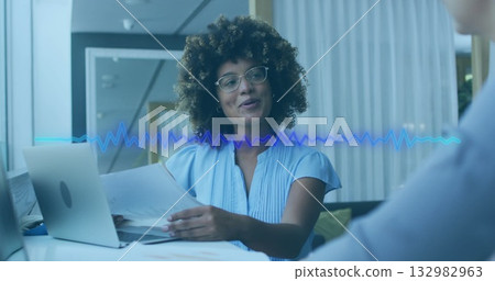 Woman in blue blouse holding papers, addressing colleague in office, with laptop and waveform Woman in blue blouse holding papers, addressing colleague in office, with laptop and waveform 132982963