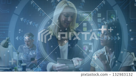 Blazer-clad blonde woman holding tablet and presenting data in open-plan office, with laptop 132983132