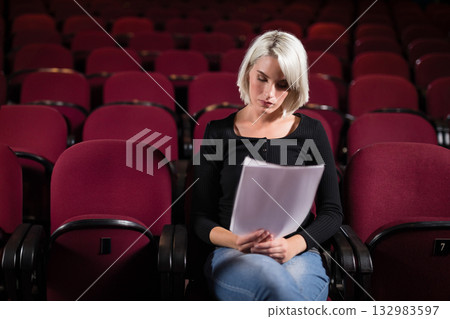 Woman wearing fitted black top reading script pages while sitting in red theater seats Woman wearing fitted black top reading script pages while sitting in red theater seats 132983597