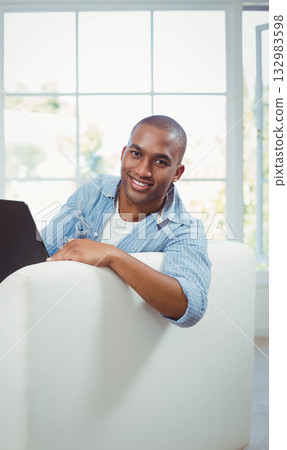 African American man wearing blue shirt, white t-shirt sitting on couch in living room using laptop 132983598