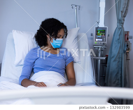 African American woman sitting up in hospital bed wearing surgical mask and monitoring IV pump 132983599