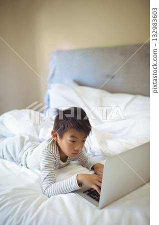 Chinese boy wearing striped pajamas lying on stomach on bed at home using silver laptop 132983603