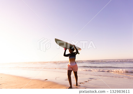 African American woman walking on beach wearing rash guard swim shorts carrying surfboard overhead African American woman walking on beach wearing rash guard swim shorts carrying surfboard overhead 132983611