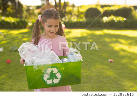 Girl wearing pink dress standing on grass holding green recycling bin with plastic bottles and bags 132983616