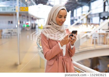 Woman wearing trench coat and headscarf leaning on railing inside terminal checking smartphone Woman wearing trench coat and headscarf leaning on railing inside terminal checking smartphone 132983621