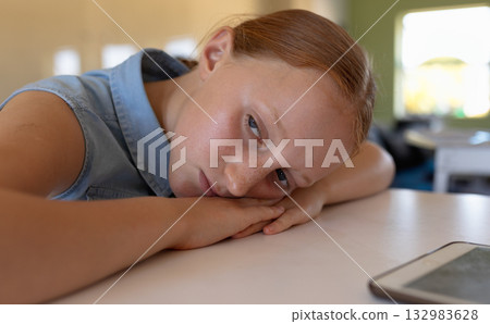 Female child resting head on arms on white desk while gazing at dark-screen tablet in classroom Female child resting head on arms on white desk while gazing at dark-screen tablet in classroom 132983628