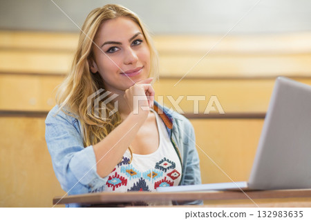 Female student sitting at lecture hall desk resting chin on hand studying with papers and laptop 132983635