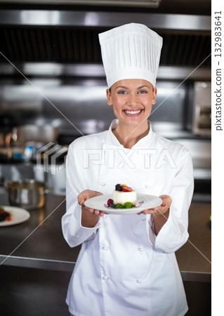 Female chef wearing coat, hat while holding dessert plate in restaurant kitchen with steel counter Female chef wearing coat, hat while holding dessert plate in restaurant kitchen with steel counter 132983641