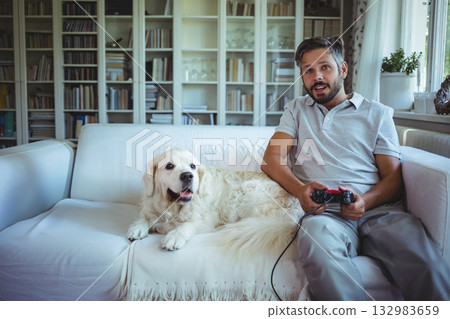 Mid adult man gaming with wired controller sitting on couch at home near bookshelf, copy space 132983659