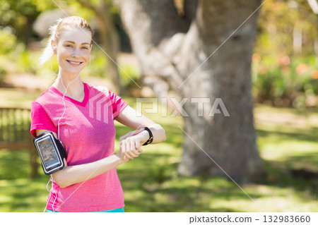 Woman checking fitness smartwatch and adjusting earbuds while holding smartphone in sunlit park 132983660