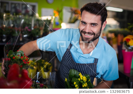 Adult male florist in apron leaning over counter in florist shop arranging colorful potted blooms Adult male florist in apron leaning over counter in florist shop arranging colorful potted blooms 132983675
