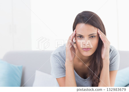 Woman in her thirties pressing temples sitting on light grey sofa in living room with cushions Woman in her thirties pressing temples sitting on light grey sofa in living room with cushions 132983679