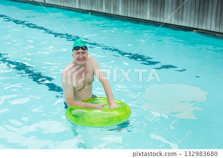 Senior white man wearing green swim cap and goggles floating in pool lanes holding lime-green ring 132983680