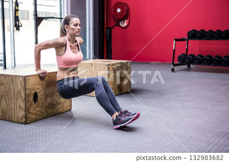 Woman in sportswear performing triceps dip gripping wooden boxes under fire hose reel in gym 132983682