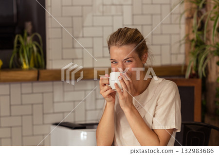 Woman sipping from white ceramic mug near mouth at cafe corner with coffee machine, potted plants Woman sipping from white ceramic mug near mouth at cafe corner with coffee machine, potted plants 132983686