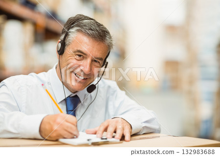 Male in business attire writing on clipboard with headset leaning on box in warehouse, copy space Male in business attire writing on clipboard with headset leaning on box in warehouse, copy space 132983688