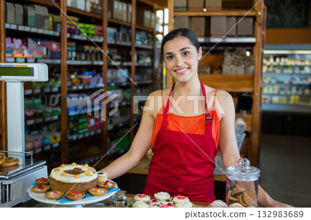 Woman wearing red apron smiling behind counter displaying frosted cake, cupcakes, cookie jar Woman wearing red apron smiling behind counter displaying frosted cake, cupcakes, cookie jar 132983689