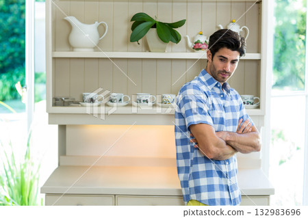 Man standing with crossed arms in kitchen nook near wooden hutch and floral teapot 132983696