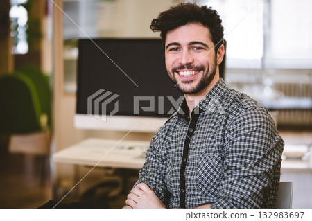 Man sitting at desk crossing arms in office workspace wearing checkered shirt with computer monitor Man sitting at desk crossing arms in office workspace wearing checkered shirt with computer monitor 132983697