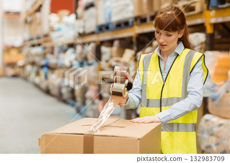 Female worker in hi-vis vest sealing cardboard box with tape dispenser in storage aisle, copy space 132983709