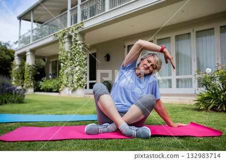 Senior woman stretching on pink, blue yoga mats in front yard with fitness tracker, copy space 132983714