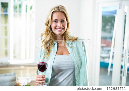 Woman wearing mint green shirt holding red wine glass and smiling in sunlit home living area Woman wearing mint green shirt holding red wine glass and smiling in sunlit home living area 132983731