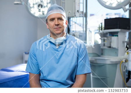 Male surgeon sitting in surgery room in scrubs beside anesthesia machine under overhead lights Male surgeon sitting in surgery room in scrubs beside anesthesia machine under overhead lights 132983733