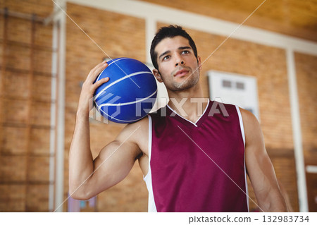 Male athlete wearing jersey holding blue basketball standing on gym court with backboard 132983734