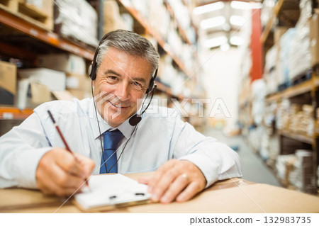 Senior man wearing tie writing on clipboard in warehouse aisle with shelving and headset microphone 132983735