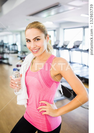 Woman in twenties smiling in gym wearing sportswear holding water bottle with towel by treadmills Woman in twenties smiling in gym wearing sportswear holding water bottle with towel by treadmills 132983739