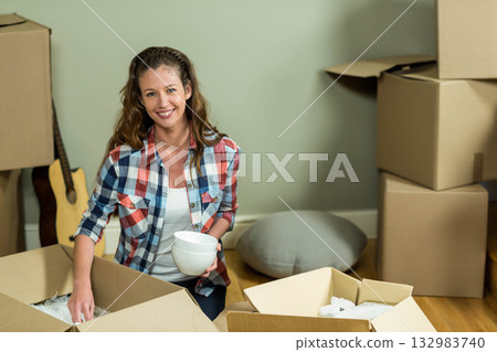 Woman kneeling on wood floor at home unpacking white ceramic bowls into cardboard boxes near guitar Woman kneeling on wood floor at home unpacking white ceramic bowls into cardboard boxes near guitar 132983740
