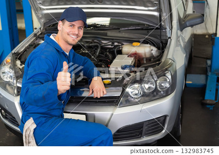 Adult male mechanic kneeling in workshop giving thumbs-up and raising silver sedan hood, copy space 132983745