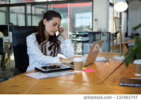 Woman talking on smartphone and typing on laptop in open-plan office with paperwork and coffee cups 132983747