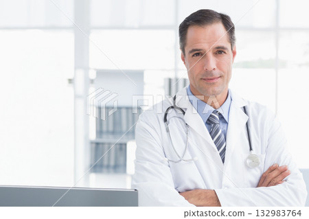 Mature male doctor standing by desk with laptop in clinic wearing coat and stethoscope, copy space 132983764