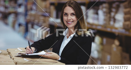 Businesswoman wearing blazer standing by metal shelves and boxes, writing on clipboard at warehouse 132983772