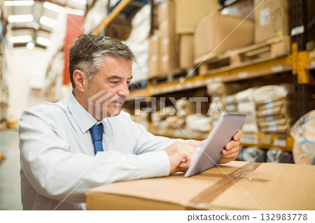 Senior white male manager in business attire standing in warehouse aisle examining tablet on box 132983778