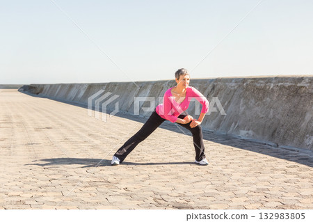 Senior woman in sportswear performing side lunge stretch on seafront walkway by seawall, copy space Senior woman in sportswear performing side lunge stretch on seafront walkway by seawall, copy space 132983805