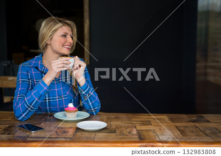 Woman in 20s sitting at cafe counter holding coffee cup with cupcake and smartphone, copy space 132983808