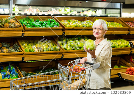Senior shopper holding green apple while pushing metal cart in grocery produce aisle, copy space 132983809